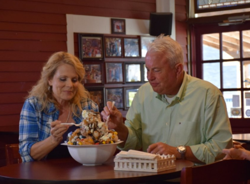 A couple enjoys a large dessert together at a restaurant, sharing bites from a bowl filled with colorful treats.