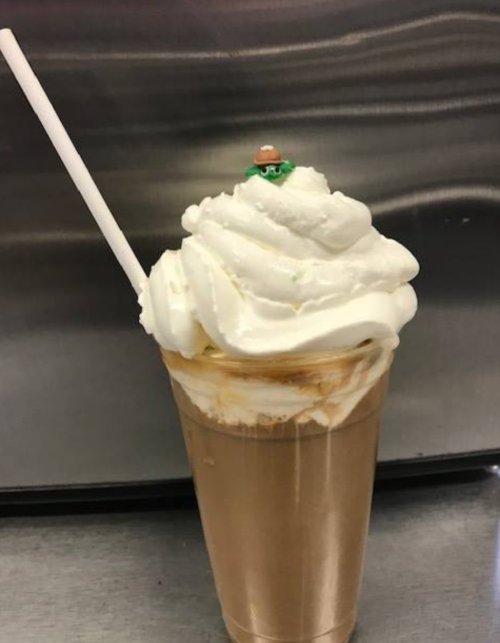 A creamy milkshake topped with whipped cream and a small decorative figure, served in a clear cup with a straw.