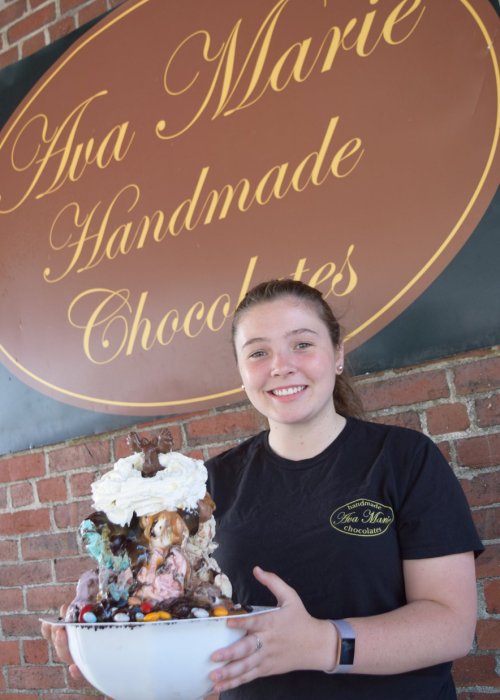 A young woman holds a large, extravagant ice cream sundae in front of a chocolate shop sign.