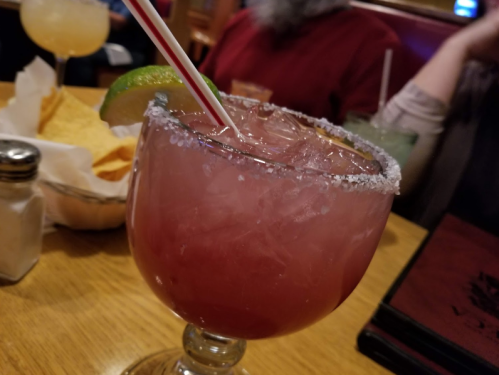 A colorful cocktail with a salted rim and a lime wedge, served on a wooden table with snacks in the background.