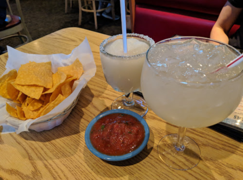 Two large margaritas and a bowl of tortilla chips with salsa on a wooden table.