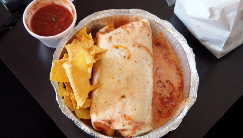 A foil container with a large burrito, tortilla chips, and a small cup of salsa on the side.
