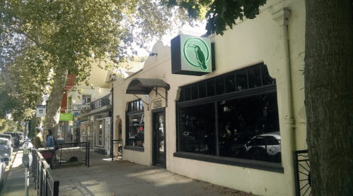 A street view of a storefront with a green sign, surrounded by trees and other shops on a sunny day.