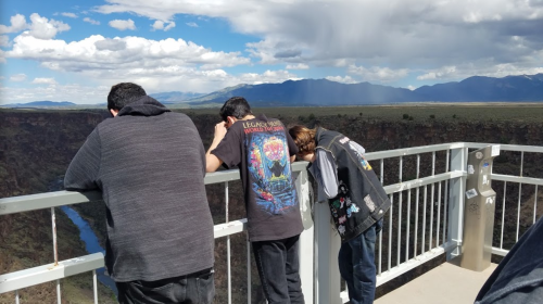 Three people lean over a railing, looking down into a canyon under a cloudy sky.