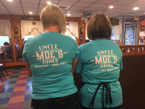 Two people wearing matching turquoise t-shirts with "Uncle Moe's Diner" printed on the back, standing in a diner.