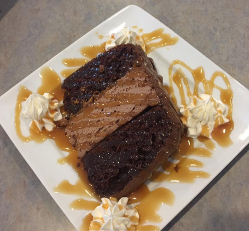 A slice of chocolate cake drizzled with caramel sauce, surrounded by whipped cream on a white plate.