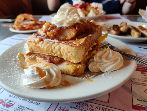 A plate of French toast topped with whipped cream, caramel, and apple filling, garnished with powdered sugar.