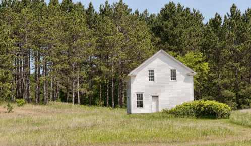A white house stands alone in a grassy field, surrounded by tall green trees under a clear blue sky.