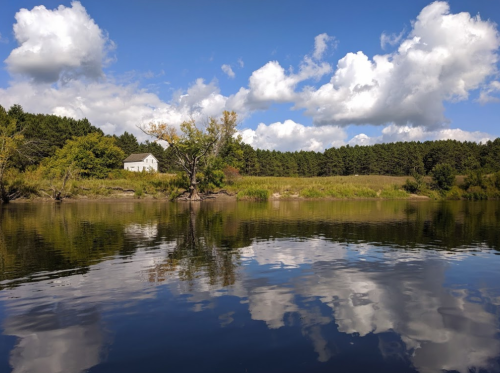 A serene landscape featuring a calm lake reflecting fluffy clouds and a white house surrounded by greenery.