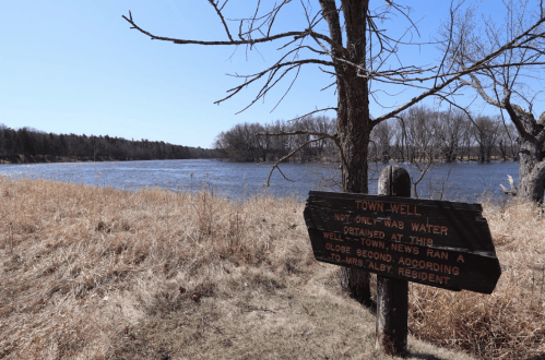 A wooden sign near a river, stating "Town Well" with a grassy area and trees in the background under a clear blue sky.