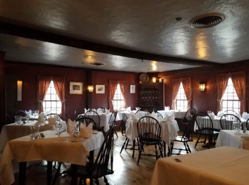 Cozy restaurant interior with wooden chairs, white tablecloths, and warm lighting from windows and wall sconces.
