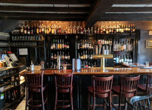 A cozy bar with a wooden counter, high stools, and shelves filled with various bottles of liquor and glassware.