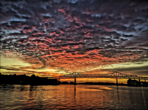 A vibrant sunset over a river, with dramatic clouds and a bridge silhouetted against the colorful sky.