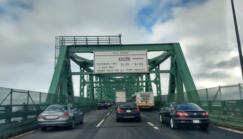 Cars driving on a green bridge with a toll rate sign displaying prices for passenger vehicles.