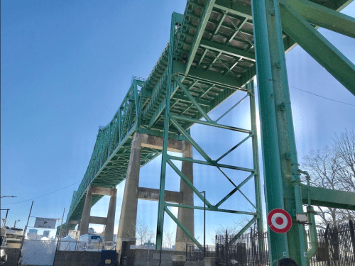 A green bridge towering above, supported by large concrete pillars, with a clear blue sky in the background.