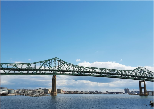 A green bridge spans over a calm river, with a clear blue sky and city skyline in the background.