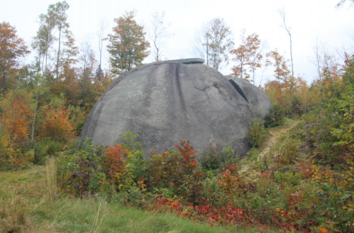 A large, smooth boulder surrounded by colorful autumn foliage and trees in a natural landscape.