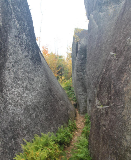 Narrow passage between large, moss-covered rocks, surrounded by greenery and autumn foliage.