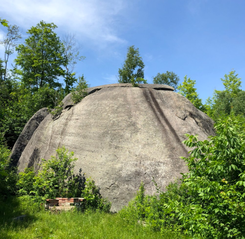 A large, rounded rock surrounded by lush green trees and shrubs under a clear blue sky.