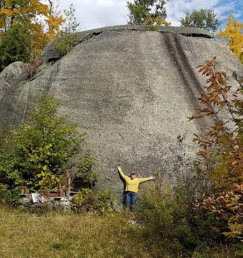 A person in a yellow sweater stands beside a large, rounded rock formation, surrounded by trees and autumn foliage.