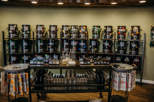 A display of various spice and seasoning jars on shelves, with a table featuring bottles and labels in a cozy shop setting.