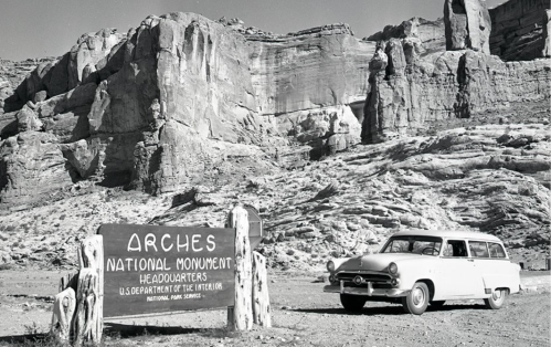 A vintage car parked near the Arches National Monument sign, surrounded by rocky cliffs in black and white.