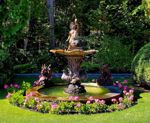 A decorative fountain with a statue surrounded by colorful flowers and lush greenery in a garden setting.