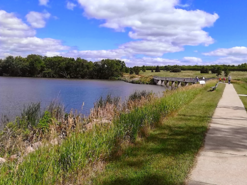 A scenic view of a riverbank with a walking path, lush greenery, and a bridge under a blue sky with fluffy clouds.