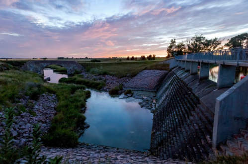 A serene landscape at sunset featuring a dam, calm water, and lush greenery under a colorful sky.