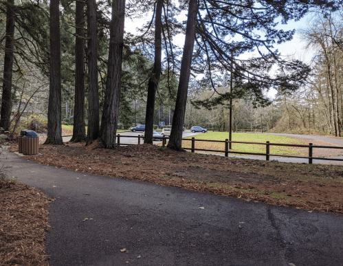 A path leads through tall trees to a parking area, with cars visible in the distance and a grassy field beyond.