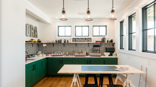 A modern kitchen with green cabinets, a large table, and decorative shelves featuring various kitchen items.