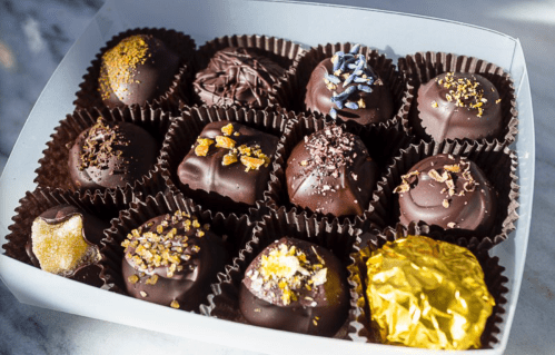A box of assorted chocolate truffles, decorated with various toppings like gold, lavender, and nuts.