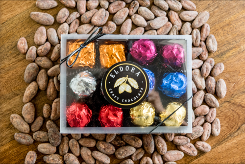 A box of assorted chocolates surrounded by cocoa beans on a wooden surface.