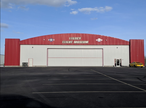 Exterior of the Legacy Flight Museum, featuring a red and white building with a large hangar door and a yellow car parked nearby.