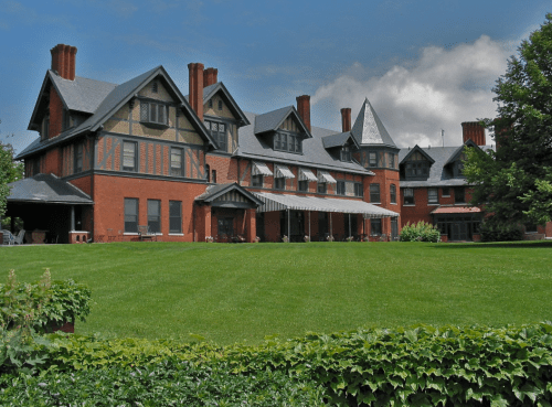 A large, historic brick mansion with multiple gables, surrounded by a lush green lawn and trees under a cloudy sky.