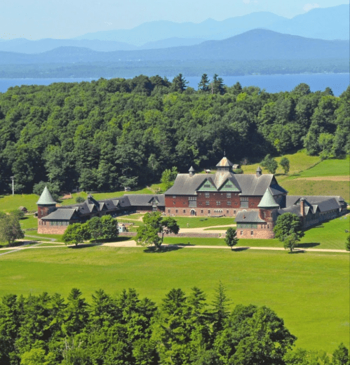Aerial view of a large, historic mansion surrounded by green fields and mountains in the background.