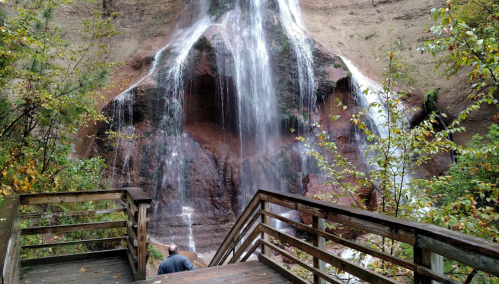 A person stands on wooden steps near a waterfall cascading down a rocky cliff, surrounded by autumn foliage.