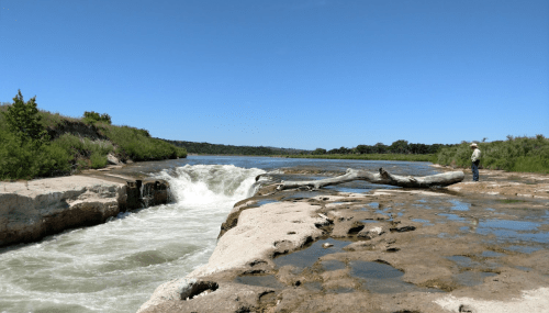 A person stands by a river with a waterfall, surrounded by greenery and rocky terrain under a clear blue sky.