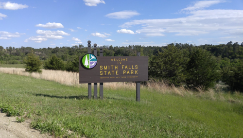 Welcome sign for Smith Falls State Park, surrounded by green grass and trees under a blue sky with clouds.