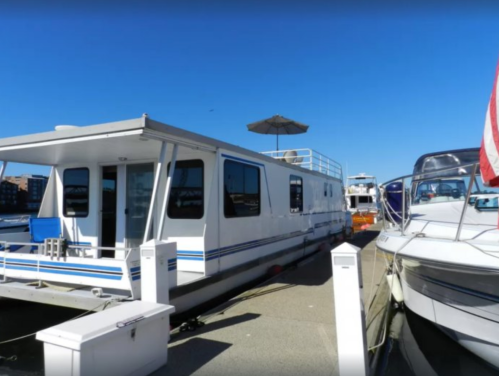 A white houseboat docked at a marina, with a parasol and nearby boats under a clear blue sky.
