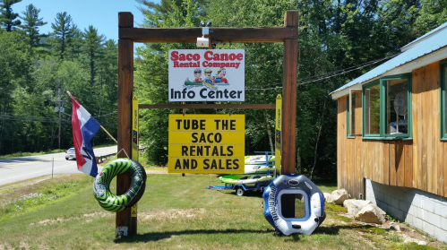 Sign for Saco Canoe Rental Company with info center, featuring tubing and rental services, surrounded by greenery.