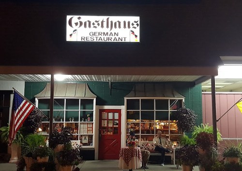 Exterior of a German restaurant named "Gasthaus," featuring a red door, decorative plants, and an American flag.