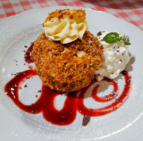A round dessert coated in nuts, topped with whipped cream, served with a berry sauce and a mint leaf on a white plate.