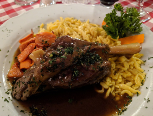 A plate of tender lamb shank with gravy, served with noodles and mixed vegetables on a checkered tablecloth.