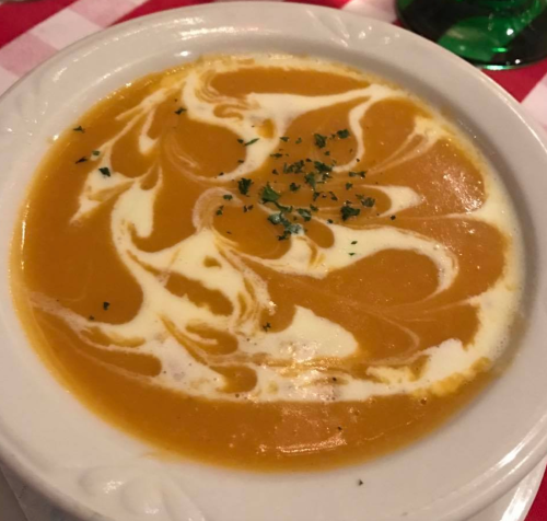 A bowl of creamy orange soup with a swirl of cream and sprinkled herbs on top, set on a checkered tablecloth.