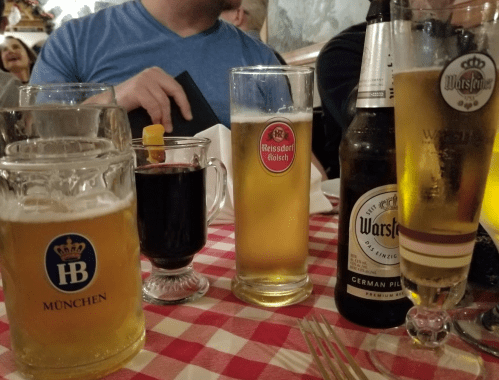 A table with various drinks: two beer glasses, a dark beverage, and a bottle, set against a checkered tablecloth.