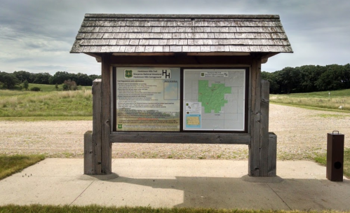 Information kiosk with a wooden roof, displaying maps and details about a natural area, set in a grassy landscape.
