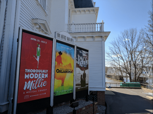 Posters for "Thoroughly Modern Millie," "Oklahoma!," and "Rags" displayed outside a theater by a river.