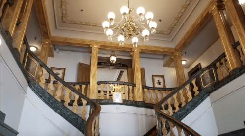 Elegant staircase with wooden railings and a chandelier, leading to a lounge area with decorative walls.