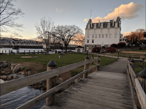 A wooden walkway leads to a historic building by the water, with trees and a bridge in the background under a colorful sky.